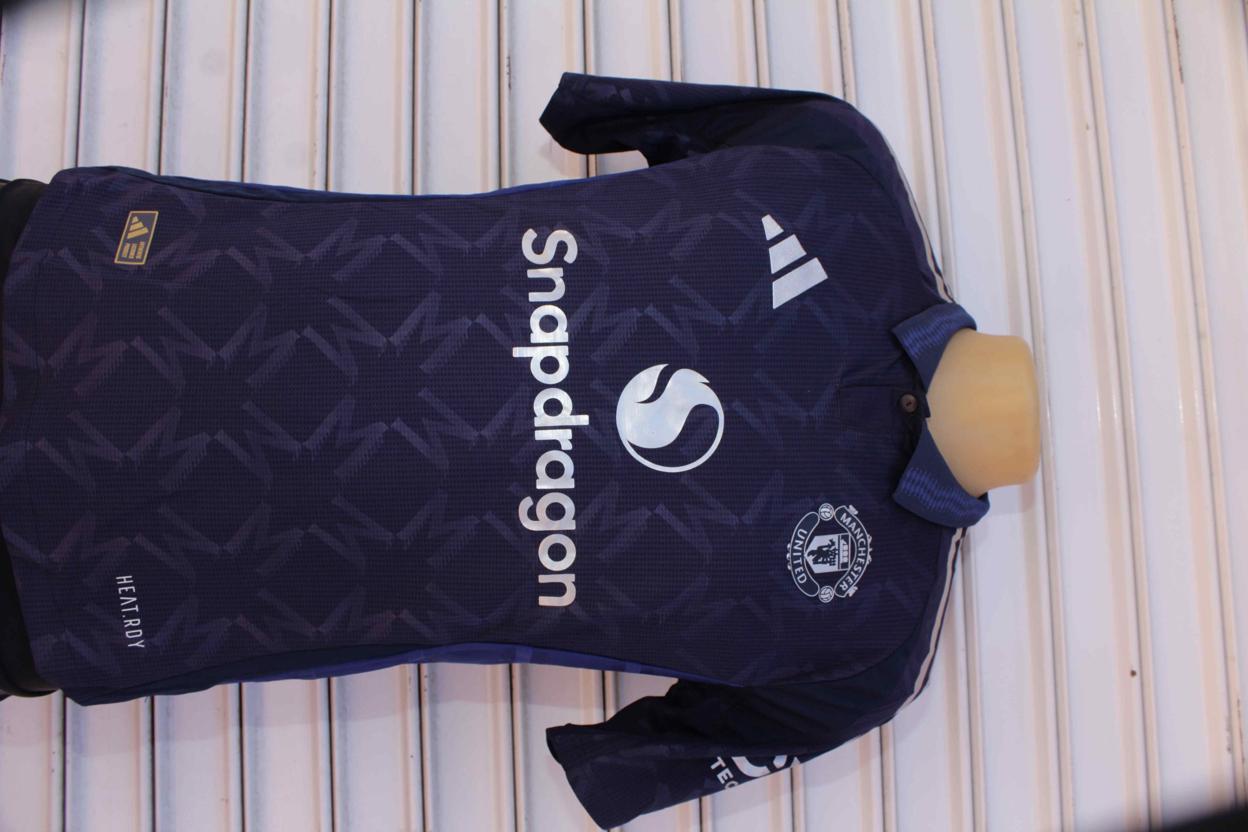 Dark blue Manchester United football jersey with geometric zigzag patterns, Adidas logo, club crest, and Snapdragon branding, displayed on a mannequin torso.