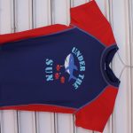 Navy blue Under the Sun Shark Graphic T-Shirt with red sleeves, side panels, and a playful shark and fish print, hanging on a wire hanger.