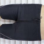 Black sports shorts with white piping along the edges and a drawstring waist, displayed on a mannequin lying on a ribbed white surface.