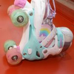Rainbow-themed roller skates with a white base, turquoise and pink accents, glitter-infused green and pink wheels, purple laces, and a rainbow star design, displayed on a red surface.