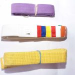 A purple belt with a metal ring, a white belt with colored rank stripes, and a yellow belt rolled with a blue rubber band, displayed on a white background.