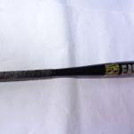 A black Easton baseball bat lying on a white surface with visible creases. The bat features a grip-wrapped handle for comfort and control. A yellow and black label near the barrel enhances its sporty look, with signs of wear on both the grip and barrel, suggesting prior use.