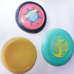 Frisbee Friends Disc Set featuring three flying discs – one red with a blue rim, one bright yellow, and one teal with a surfing-themed design, placed on a white surface.
