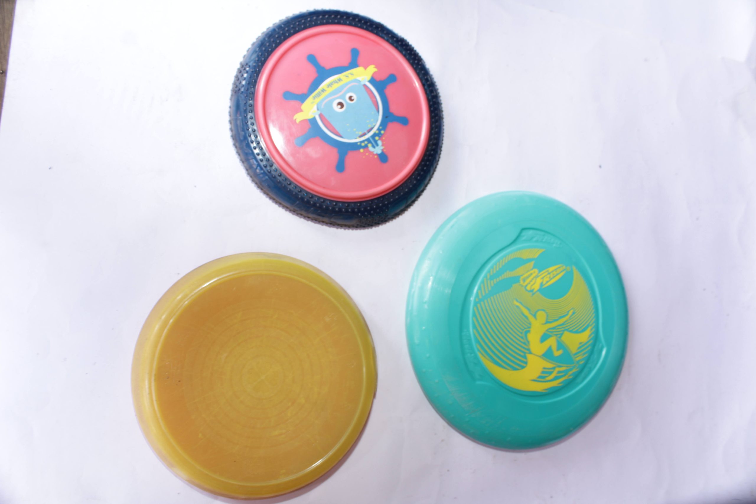 Frisbee Friends Disc Set featuring three flying discs – one red with a blue rim, one bright yellow, and one teal with a surfing-themed design, placed on a white surface.