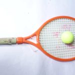 Orange tennis racket with green butterfly and flower grip, red markings on strings, and a yellow ball, placed on a white background.