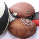 A black and white rugby ball with patent markings, a brown American football with a barcode sticker, a worn leather ball with faded text, and a textured black and red ball, displayed together.