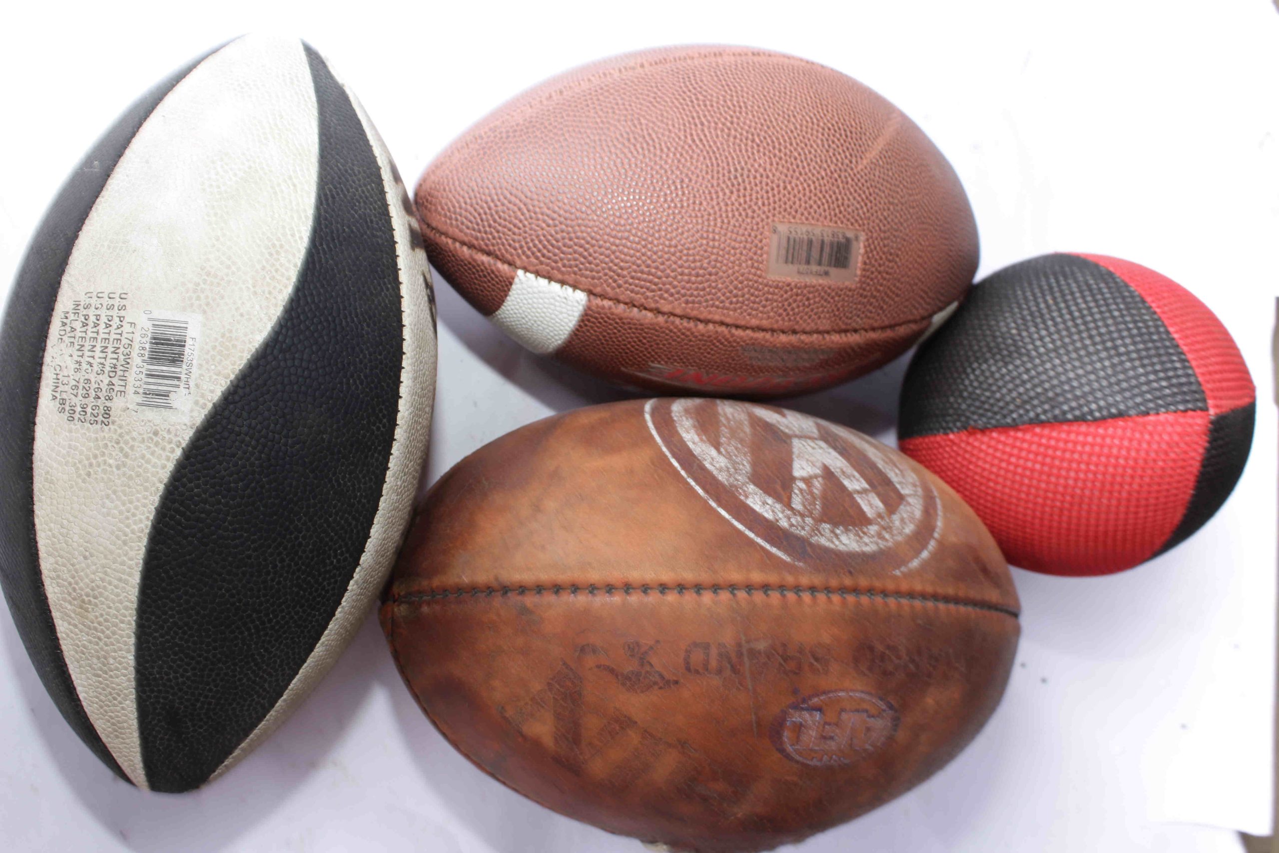 A black and white rugby ball with patent markings, a brown American football with a barcode sticker, a worn leather ball with faded text, and a textured black and red ball, displayed together.
